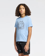 A curly-haired person stands against a plain background wearing Psycho Bunny's eco-friendly KIDS MELBOURNE GRAPHIC TEE in light blue, made from Peruvian Pima cotton, featuring a bunny and crossbones design. They also wear black shorts with a small repeating pattern.