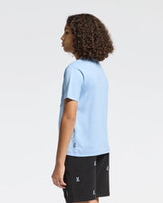 A person with curly hair stands sideways, showcasing a light blue Psycho Bunny KIDS MELBOURNE GRAPHIC TEE - B0U410F200 made from eco-friendly Peruvian Pima cotton, paired with black shorts featuring small white designs against a plain white backdrop.