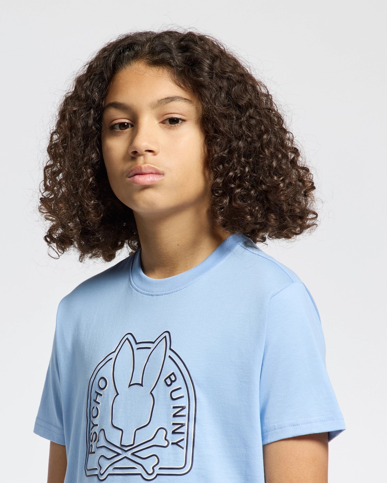 A person with curly hair is wearing a light blue KIDS MELBOURNE GRAPHIC TEE - B0U410F200 made from eco-friendly Peruvian Pima cotton. It showcases a bunny with crossbones and "Psycho Bunny" text on the front against a plain white backdrop.