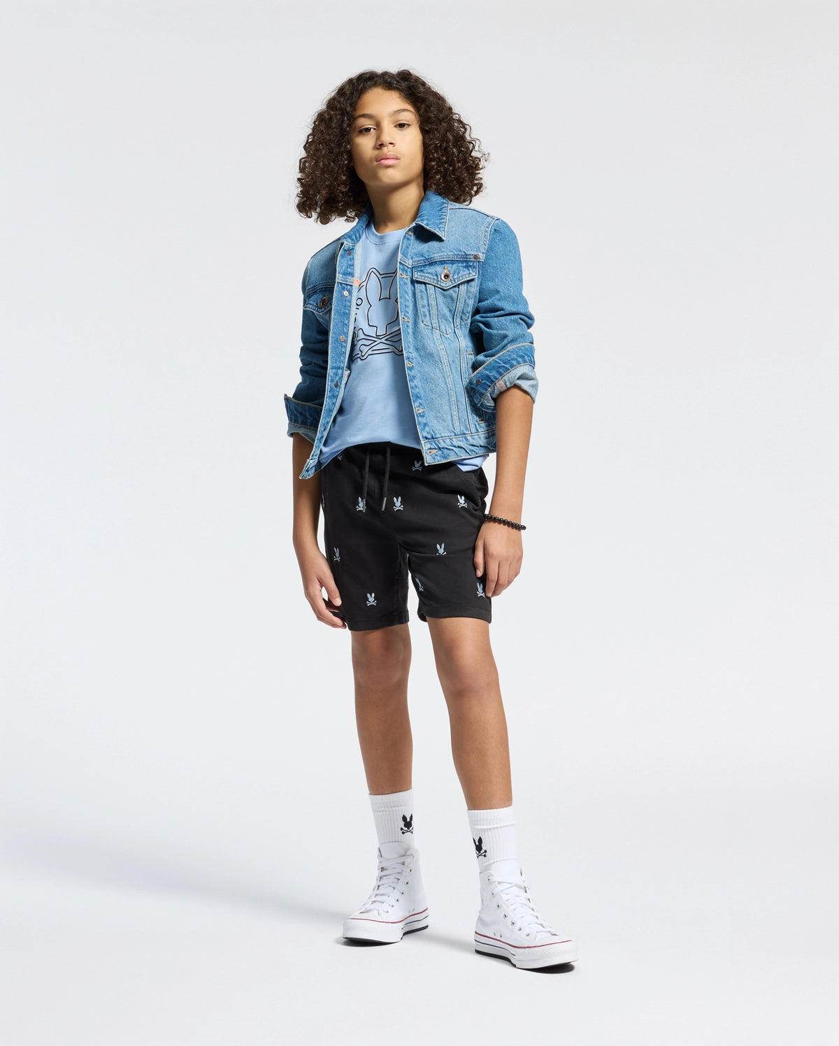 A young person with curly hair poses confidently against a light background, styled in a denim jacket over the eco-friendly KIDS MELBOURNE GRAPHIC TEE by Psycho Bunny and black shorts with white patterns, finished with white socks and high-top sneakers.