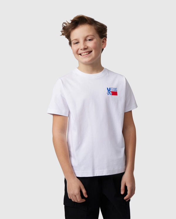 A smiling boy is wearing a Psycho Bunny KIDS TEXAS CITY TEE - B0U553B2PC made of Peruvian Pima cotton, featuring a small blue and red graphic on the chest. He's paired it with black pants against a light gray background.