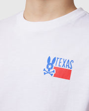 Close-up of a person wearing the Psycho Bunny KIDS TEXAS CITY TEE in white, made from luxurious Peruvian Pima cotton. The vibrant graphic design features a stylized rabbit with bone-like ears and "TEXAS" above a red rectangle displaying the Texas flag pattern.