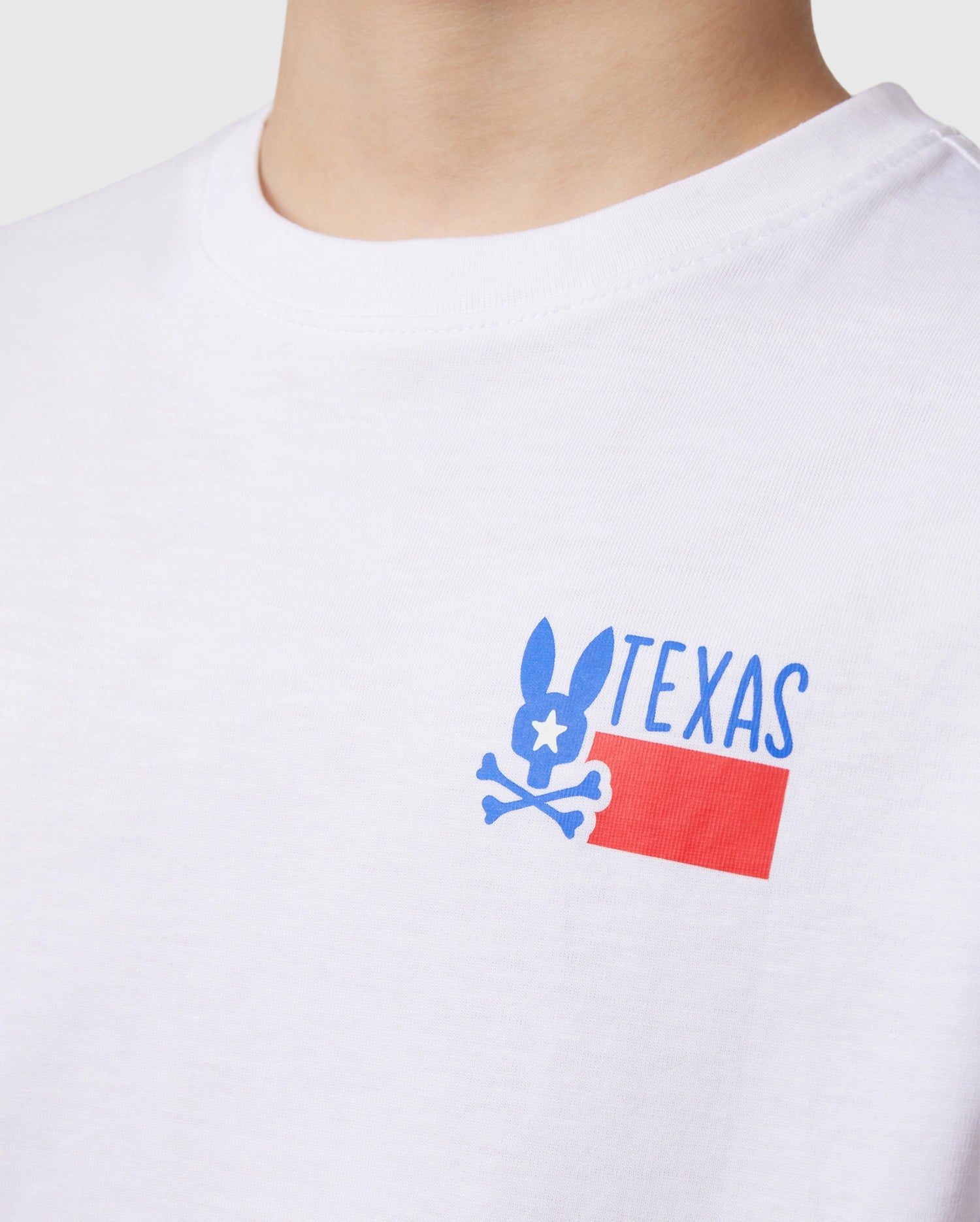Close-up of a person wearing the Psycho Bunny KIDS TEXAS CITY TEE in white, made from luxurious Peruvian Pima cotton. The vibrant graphic design features a stylized rabbit with bone-like ears and "TEXAS" above a red rectangle displaying the Texas flag pattern.