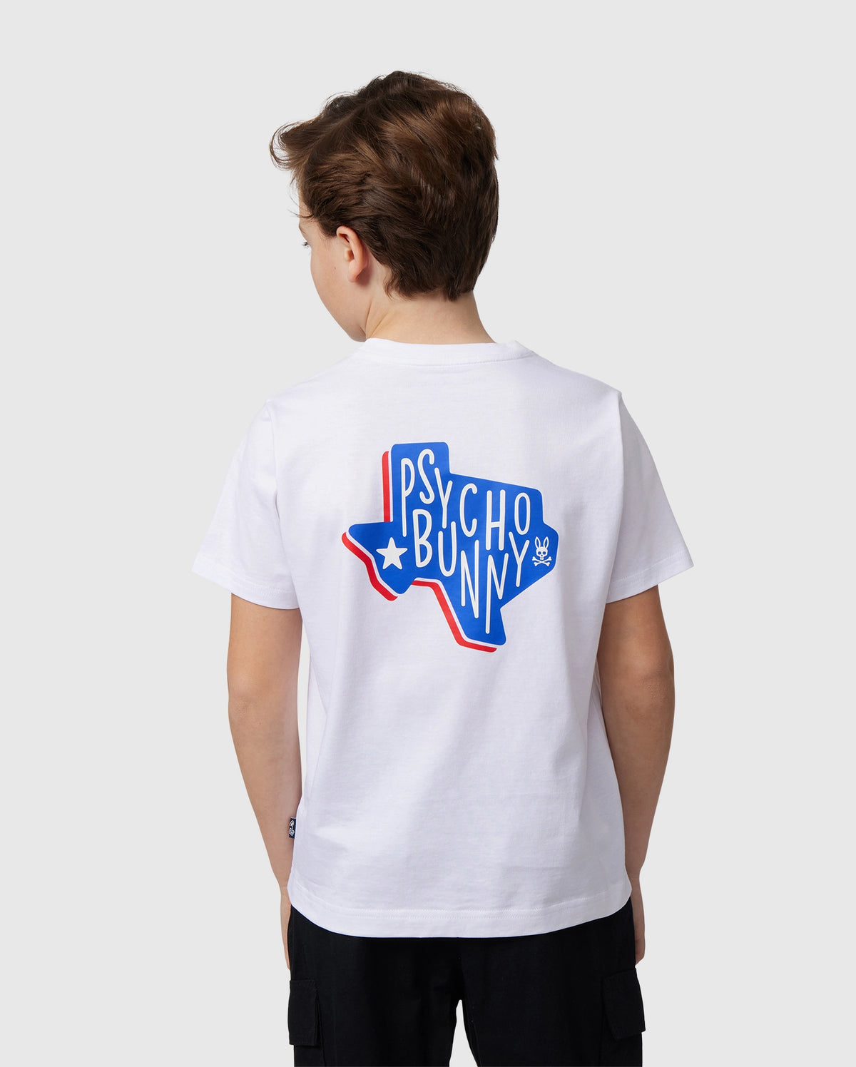 The boy dons a white KIDS TEXAS CITY TEE - B0U553B2PC by Psycho Bunny, made from soft Peruvian Pima cotton. A vibrant graphic shows "Psycho Bunny" in bold blue and red on the back. His short brown hair and gaze away from the camera add mystery to his style.