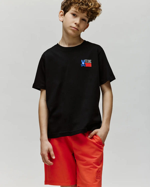 A boy with curly hair wears the Psycho Bunny KIDS TEXAS TEE (B0U553W1PC) in black, featuring a small "TEXAS" graphic, and red shorts. He stands against a light gray background, looking at the camera with a relaxed expression.