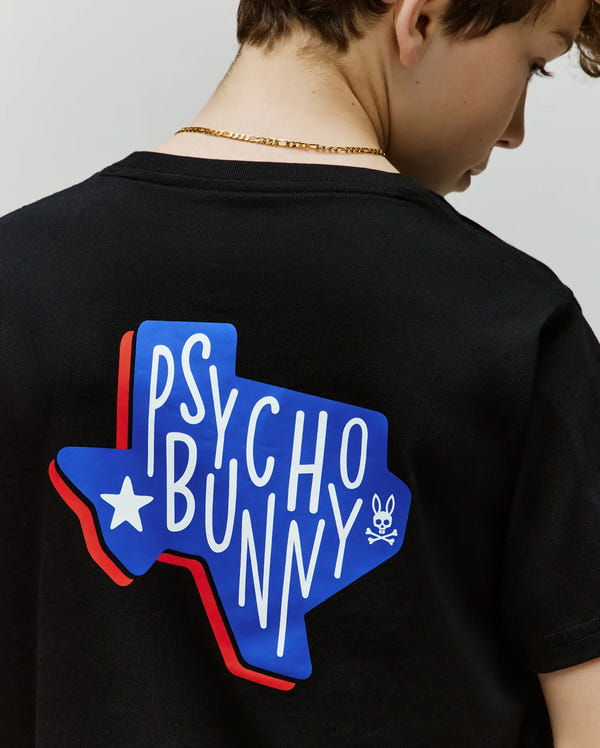 A child wears the Psycho Bunny KIDS TEXAS TEE (B0U553W1PC), a black Pima cotton shirt with a blue and red Texas outline, "Psycho Bunny" in bold white letters, and a prominent Bunny skull graphic on the back.