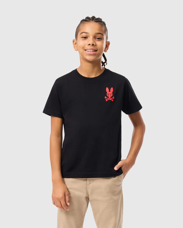 A young person with braided hair smiles while wearing a stylish Psycho Bunny KIDS NEW YORK CITY TEE in black, crafted from soft Pima cotton and featuring a red emblem of a bunny and crossbones. They pair it with beige pants, one hand casually in their pocket, standing against a plain white background.