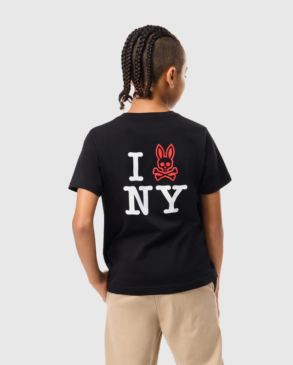 A person with braided hair is wearing the KIDS NEW YORK CITY TEE - B0U554W1PC by Psycho Bunny, made from black Pima cotton and featuring the design "I (red bunny face) NY" on the back. Their hands are tucked into the pockets of beige pants as they look away against a plain gray background.
