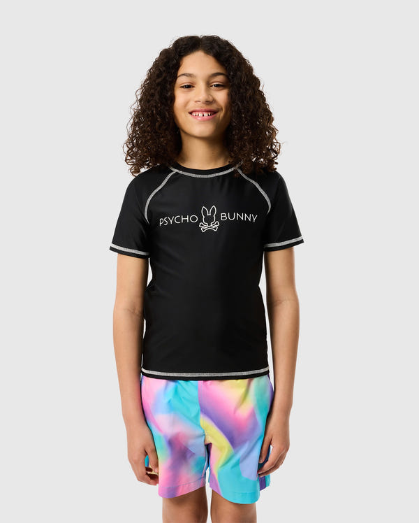 A young person with curly hair is smiling and standing against a plain background. They are wearing a black "KIDS SHORT SLEEVE ANTI-UV RASHGUARD - B0U558C200" by Psycho Bunny with an outline of a bunny on the chest, which doubles as a kids rash guard providing UPF 50+ protection, and colorful, tie-dye shorts in shades of blue, pink, green, and yellow.