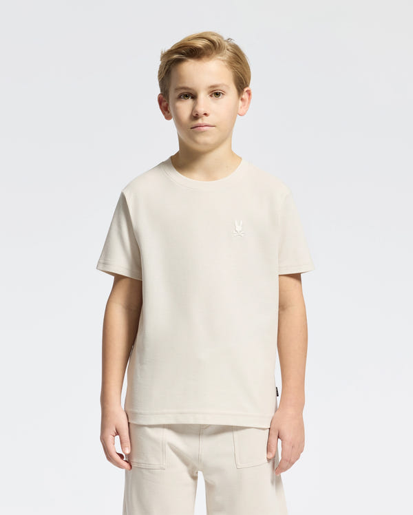 A young boy stands facing forward in a light beige KIDS TAYLOR PIQUE TEE by Psycho Bunny, paired with matching shorts. The Pima cotton tee has an embroidered Bunny logo on the chest against a plain white background.