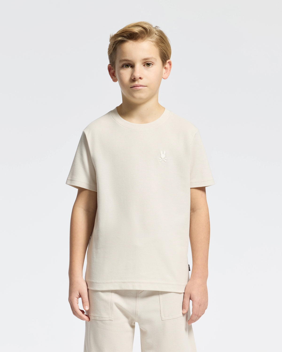 A young boy stands facing forward in a light beige KIDS TAYLOR PIQUE TEE by Psycho Bunny, paired with matching shorts. The Pima cotton tee has an embroidered Bunny logo on the chest against a plain white background.