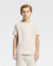 A young boy stands facing forward in a light beige KIDS TAYLOR PIQUE TEE by Psycho Bunny, paired with matching shorts. The Pima cotton tee has an embroidered Bunny logo on the chest against a plain white background.