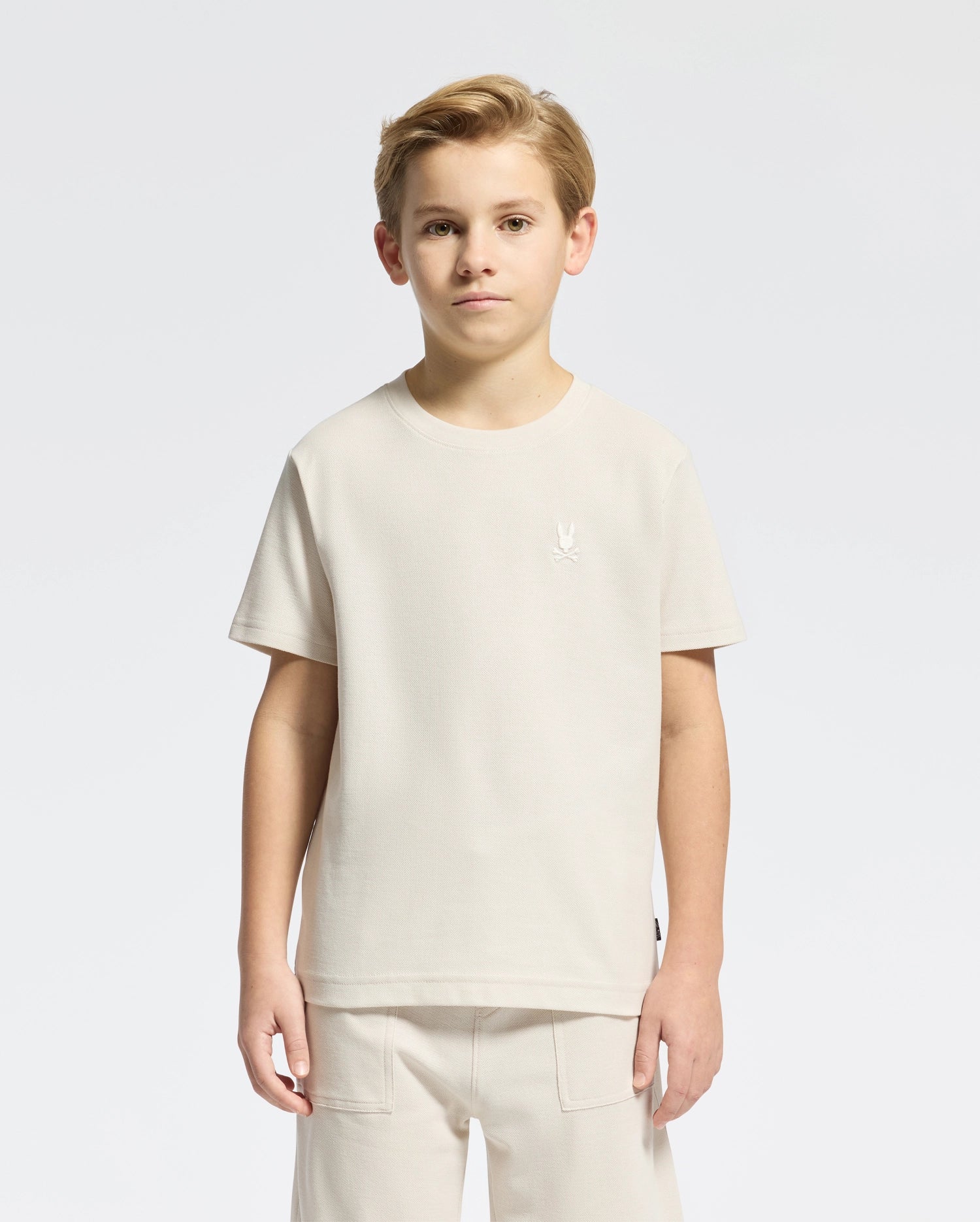 A young boy stands facing forward in a light beige KIDS TAYLOR PIQUE TEE by Psycho Bunny, paired with matching shorts. The Pima cotton tee has an embroidered Bunny logo on the chest against a plain white background.