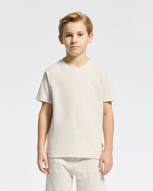A young boy stands facing forward in a light beige KIDS TAYLOR PIQUE TEE by Psycho Bunny, paired with matching shorts. The Pima cotton tee has an embroidered Bunny logo on the chest against a plain white background.