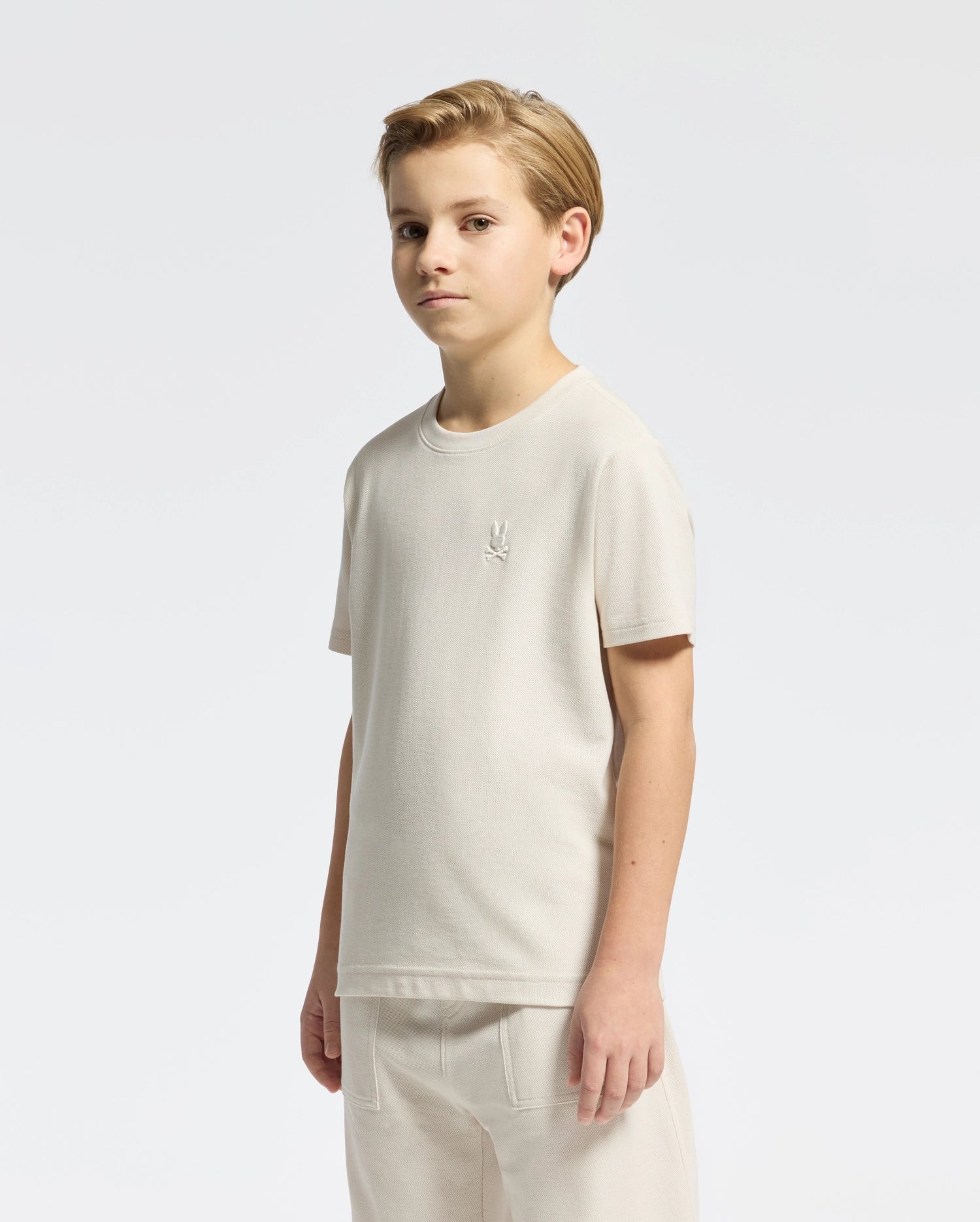 A person with short, light hair is wearing the Psycho Bunny KIDS TAYLOR PIQUE TEE (B0U579F200), a regular fit, light-colored Pima cotton tee featuring an embroidered Bunny logo on the upper left side. They are also wearing lightweight Pima cotton pants, all set against a plain white background.