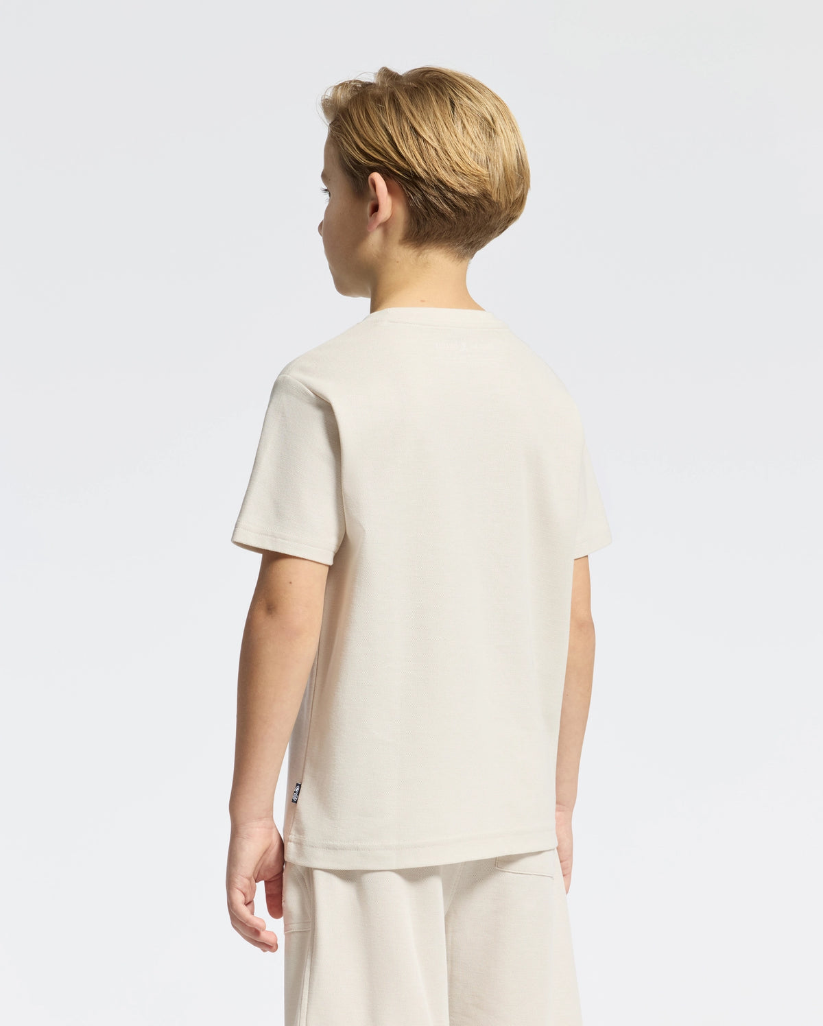 A young blonde boy stands facing away wearing the Psycho Bunny KIDS TAYLOR PIQUE TEE, B0U579F200, a regular-fit cream Pima cotton shirt paired with shorts on a plain white background.