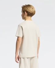 A young blonde boy stands facing away wearing the Psycho Bunny KIDS TAYLOR PIQUE TEE, B0U579F200, a regular-fit cream Pima cotton shirt paired with shorts on a plain white background.