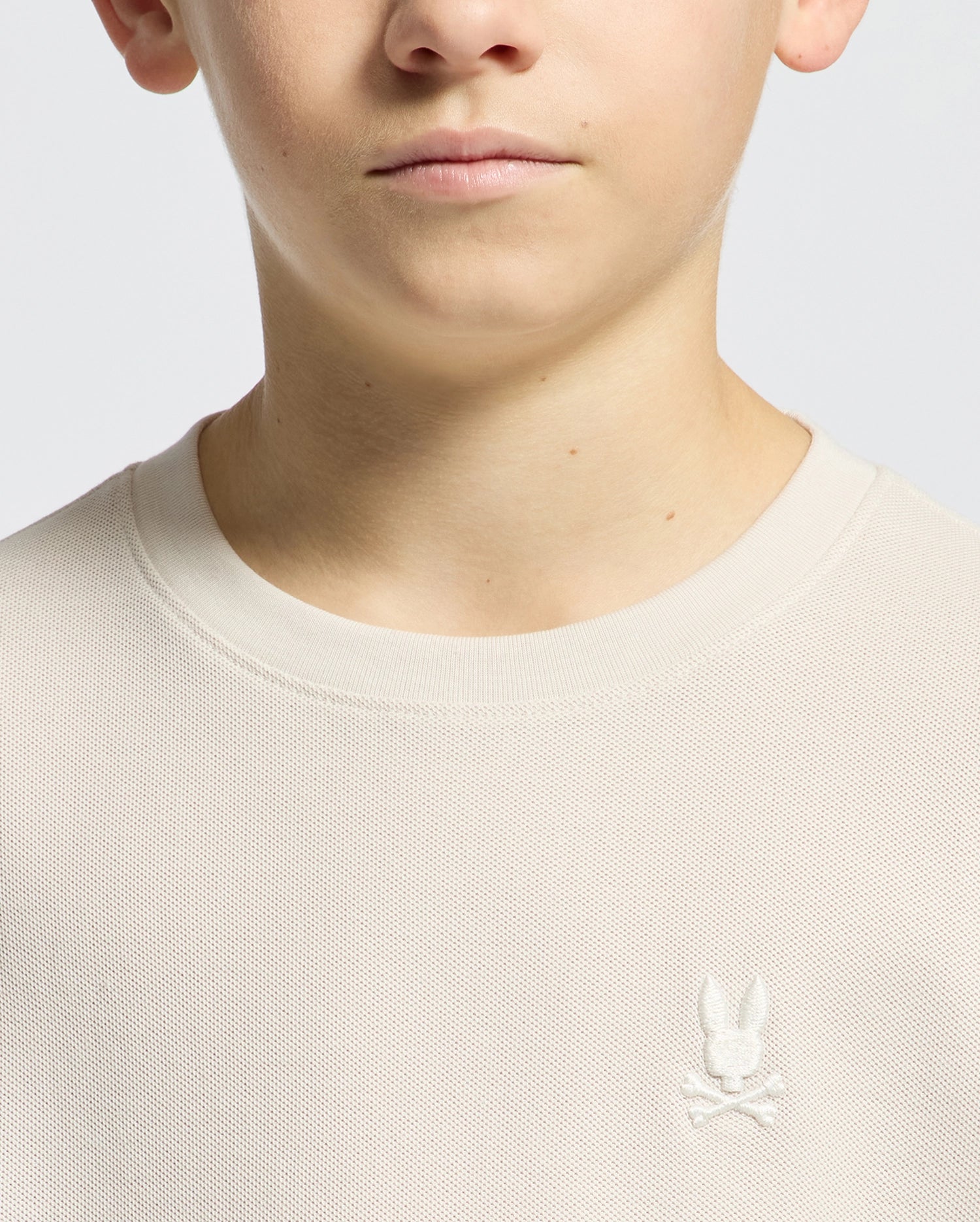 A person wears a KIDS TAYLOR PIQUE TEE by Psycho Bunny, in light beige with a regular fit round-neck design, made of lightweight Pima cotton. The chest features an embroidered Bunny emblem with crossed bones against a plain gray background.