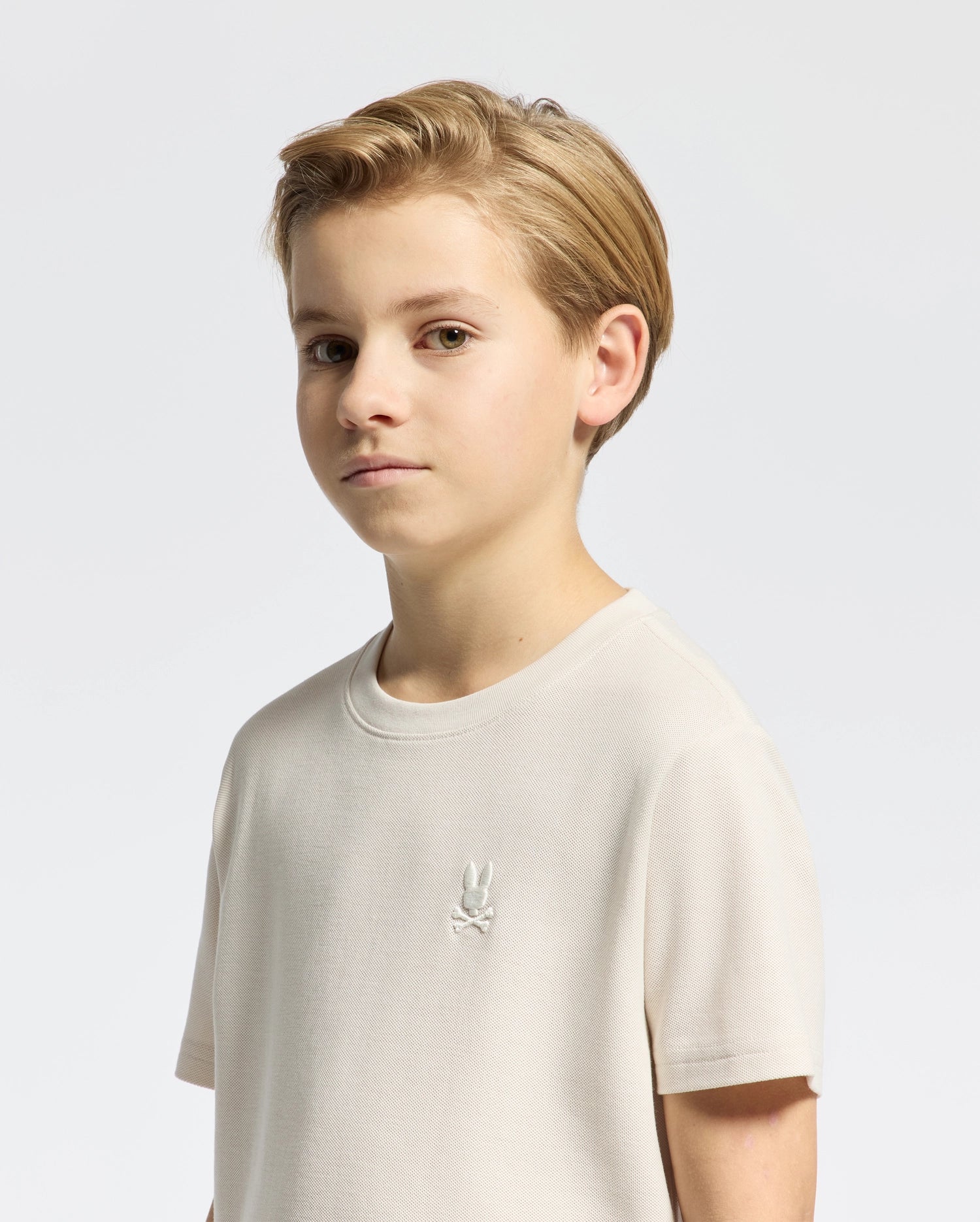 A young person with short, light hair wears the KIDS TAYLOR PIQUE TEE by Psycho Bunny. The regular fit, light-colored Pima cotton shirt features an embroidered Bunny emblem on the chest. They look toward the camera against a plain white background.