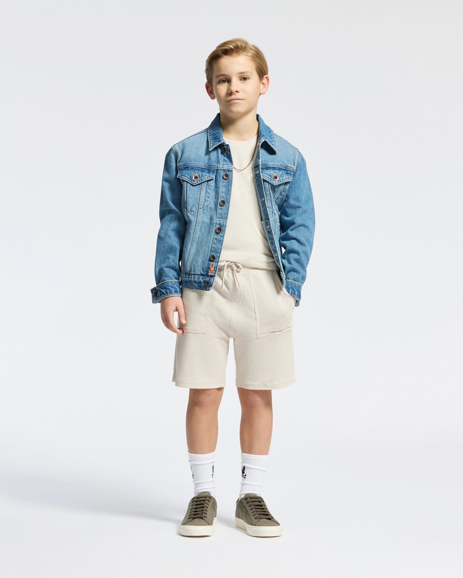 A young boy stands against a plain backdrop, wearing the KIDS TAYLOR PIQUE TEE - B0U579F200 by Psycho Bunny, styled with a blue denim jacket and matching beige shorts. He sports white socks and green sneakers, hands relaxed by his sides, with an embroidered Bunny subtly adorning the shirt corner.