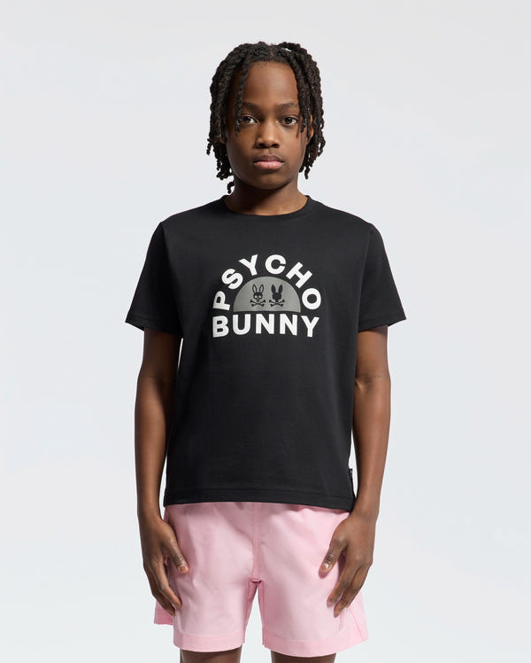 A person in a KIDS GOLDIE GRAPHIC TEE by Psycho Bunny, crafted from soft Peruvian Pima cotton, stands with hands in pink shorts pockets against a plain white background. They have shoulder-length braided hair and a neutral expression.