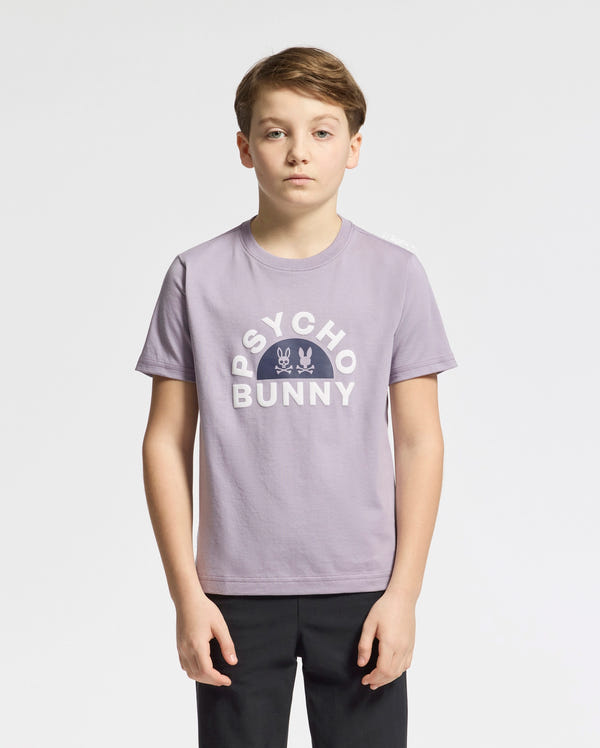 A young person in a light purple KIDS GOLDIE GRAPHIC TEE - B0U606F200 from Psycho Bunny, made of luxurious Peruvian Pima cotton, is set against a white background. The tee features a stylized bunny graphic and text. They have short, light brown hair and wear black pants.