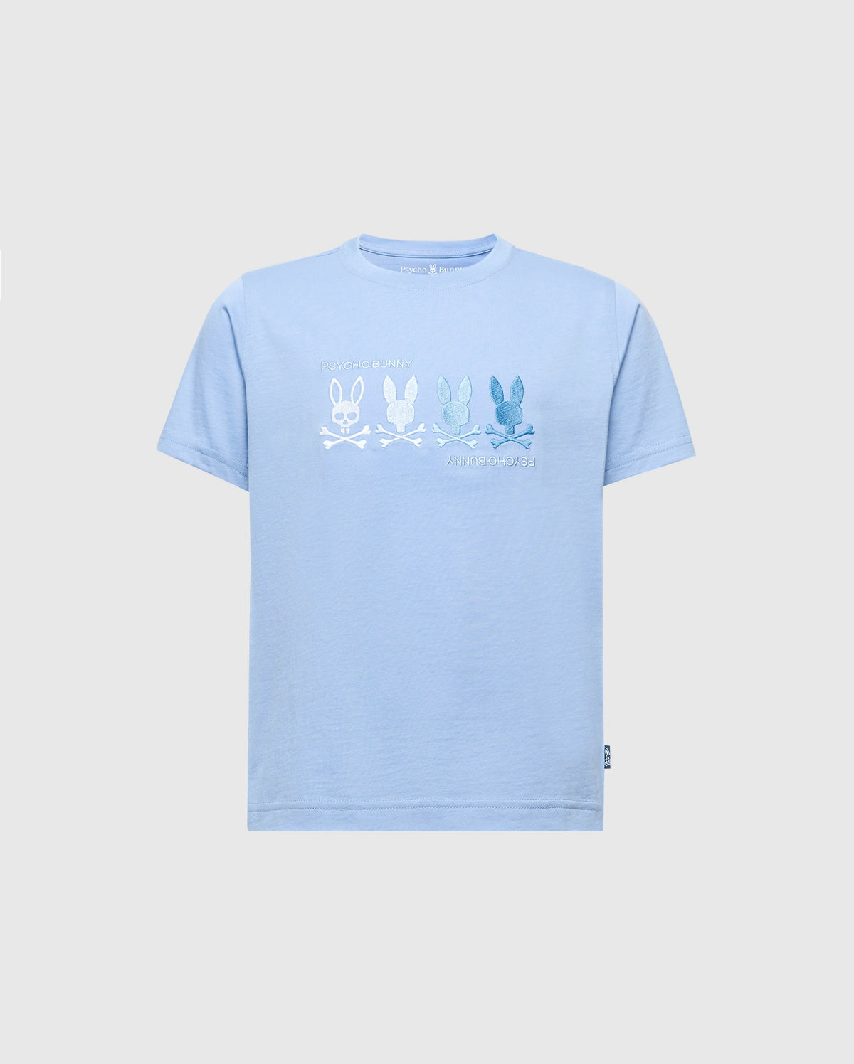 The KIDS OCEANIA EMBROIDERED GRAPHIC TEE by Psycho Bunny, product B0U607F200, features retro light blue Pima cotton with embroidered bunny skull and crossbones in varying shades. Blue bunnies are on the right and a white bunny on the left. A small label decorates the bottom left hem.