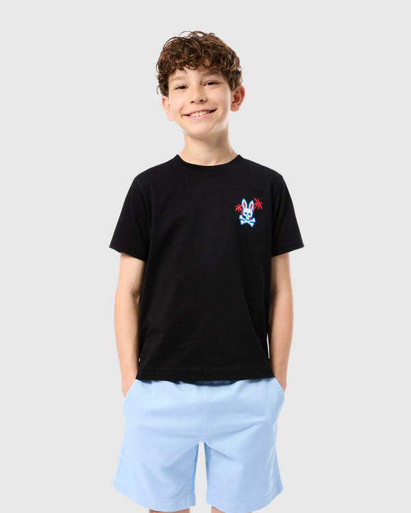A smiling child with short curly hair is wearing a Psycho Bunny KIDS LOS ANGELES CITY TEE - B0U861A2PC made from Peruvian Pima cotton, featuring a small colorful graphic design on the left side of the chest and light blue shorts. The child has their hands in their pockets, standing against a plain white background.