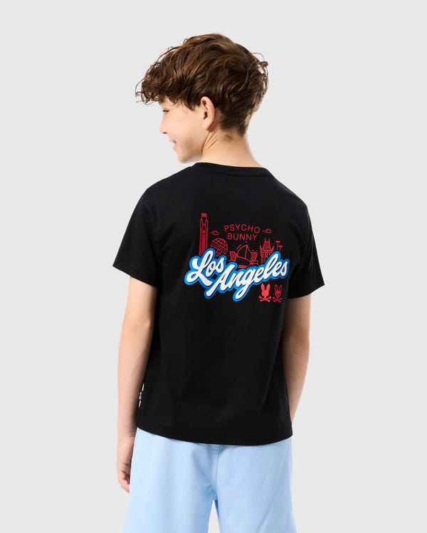 A child with curly hair, seen from behind, wears the KIDS LOS ANGELES CITY TEE - B0U861A2PC by Psycho Bunny. The soft black Peruvian Pima cotton tee showcases a vibrant graphic design of buildings and the text "Los Angeles" alongside "Psycho Bunny." Light blue shorts complete the look against a gray background.