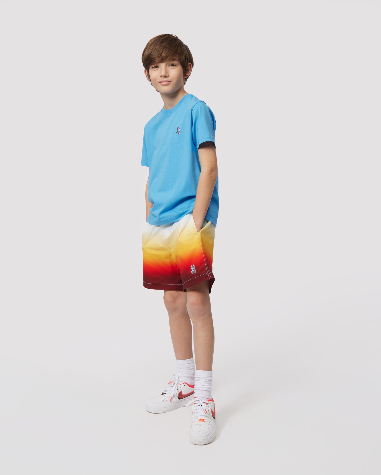 A young boy in a blue t-shirt and vibrant print Psycho Bunny kids' swim trunk (KIDS GUY SWIM TRUNK - B0W114Y1PO) stands against a white background, looking at the camera with a slight smile, wearing white sneakers.