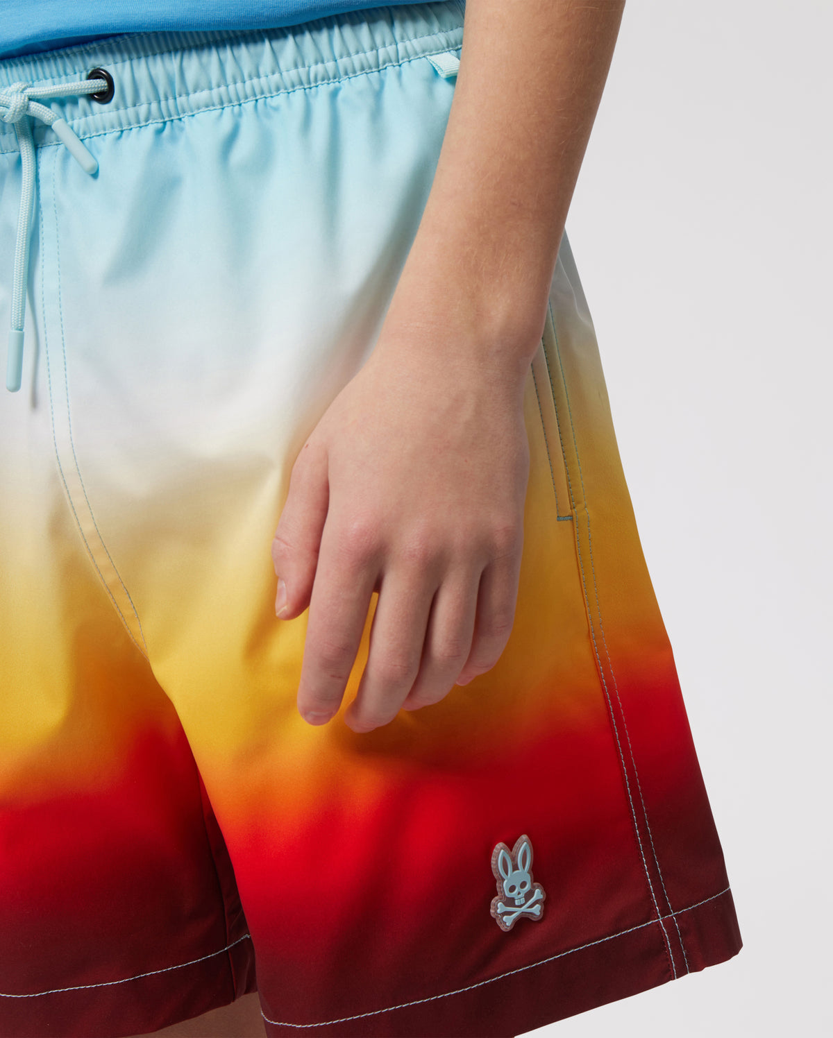 Close-up of a person wearing vibrant print Psycho Bunny kids' swim trunk ranging from sky blue to fiery red, with a small silver bunny logo on the lower left leg.
