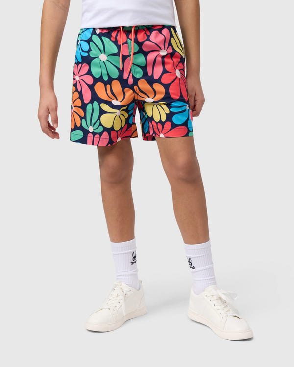 A person is wearing the KIDS MENTZ SWIM TRUNK - B0W320B2SW from Psycho Bunny, featuring a vibrant, quick-dry fabric with a bold, abstract floral pattern in shades of red, yellow, green, blue, and pink. They are also sporting white socks and white sneakers while standing against a plain gray background. Only their lower body is visible.