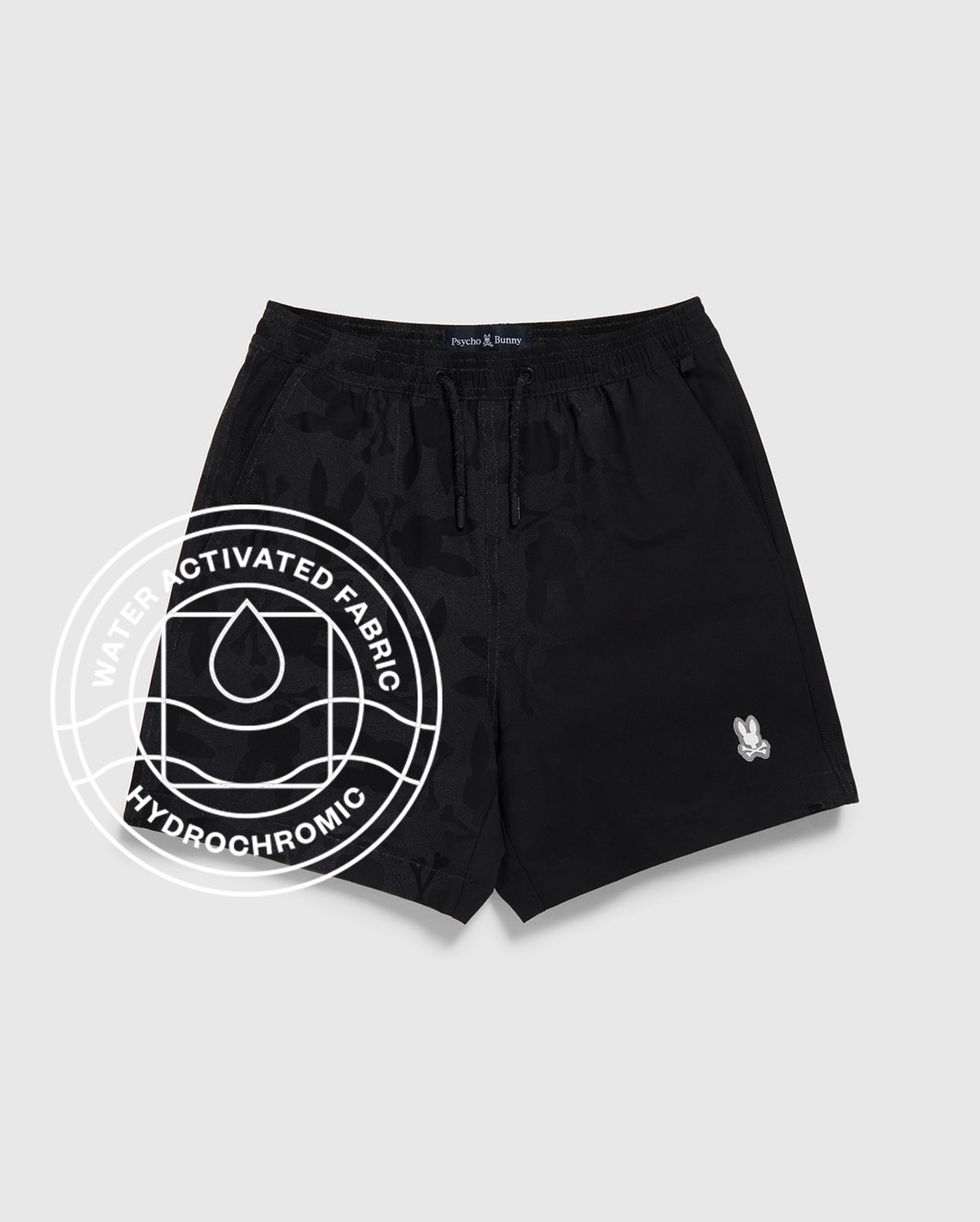 The KIDS PARKER HYDROCHROMIC SWIM TRUNK by Psycho Bunny in black features a subtle camo hydrochromatic pattern, quick-dry fabric, drawstring waistband, and a white logo patch. An overlayed graphic highlights its water-activated fabric. Displayed on light grey.