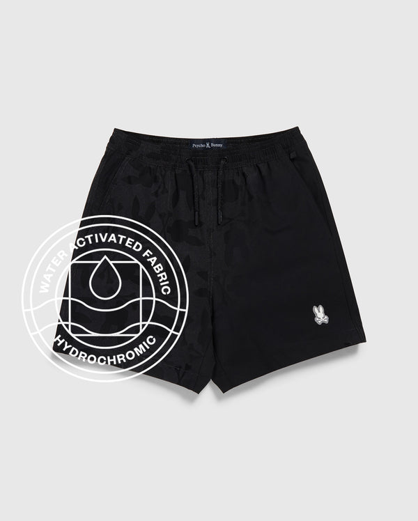 The KIDS PARKER HYDROCHROMIC SWIM TRUNK by Psycho Bunny in black features a subtle camo hydrochromatic pattern, quick-dry fabric, drawstring waistband, and a white logo patch. An overlayed graphic highlights its water-activated fabric. Displayed on light grey.