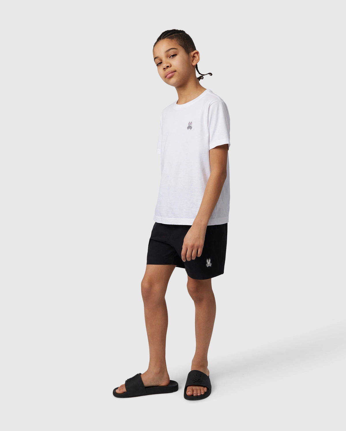 A young person stands casually wearing a white t-shirt, Psycho Bunny KIDS MALTA HYDROCHROMIC SWIM TRUNK - B0W321B2SW, and black sandals, facing slightly to their left, with a neutral expression and hands by their sides.