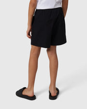 A person standing, wearing black Psycho Bunny swim trunks and black slip-on shoes, viewed from behind. The focus is on the clothing, specifically the swim trunks.