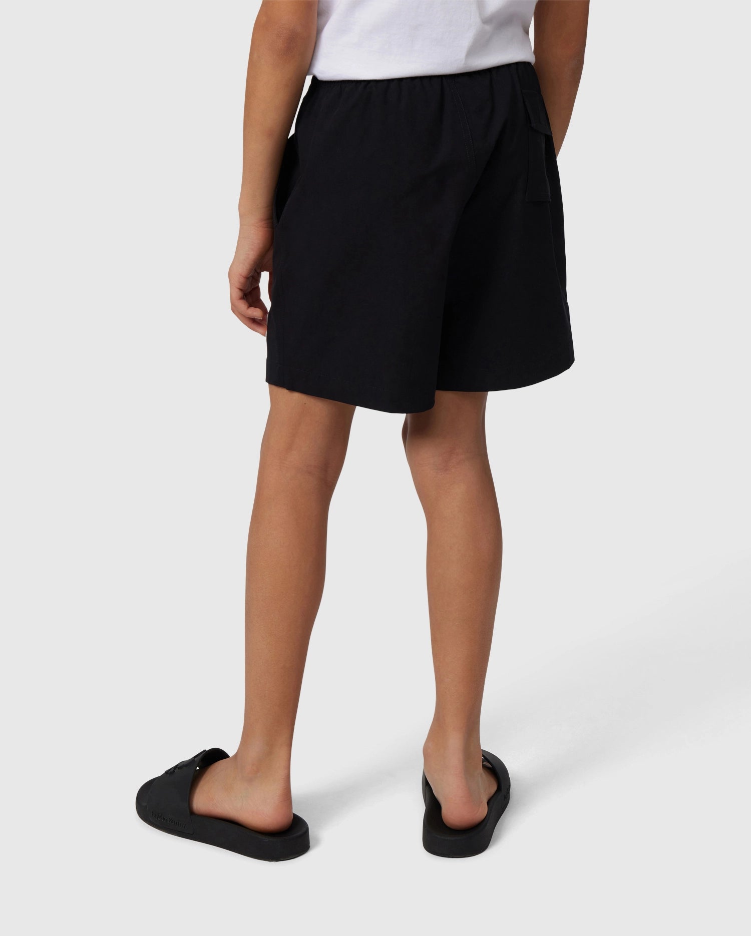 A person standing, wearing black Psycho Bunny swim trunks and black slip-on shoes, viewed from behind. The focus is on the clothing, specifically the swim trunks.