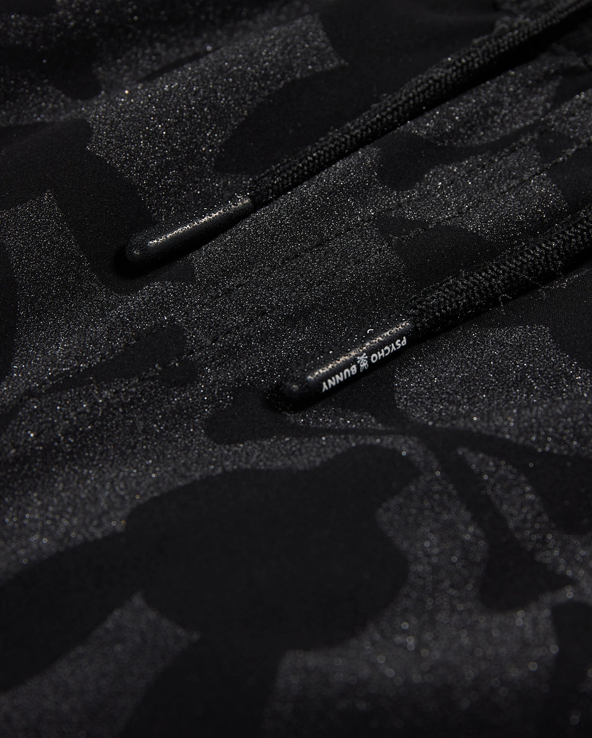 Close-up of black polyester mechanical stretch fabric with a subtle camo pattern and glossy accents, featuring drawstrings with visible Psycho Bunny branding.