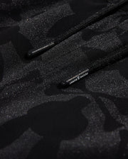 Close-up of black polyester mechanical stretch fabric with a subtle camo pattern and glossy accents, featuring drawstrings with visible Psycho Bunny branding.