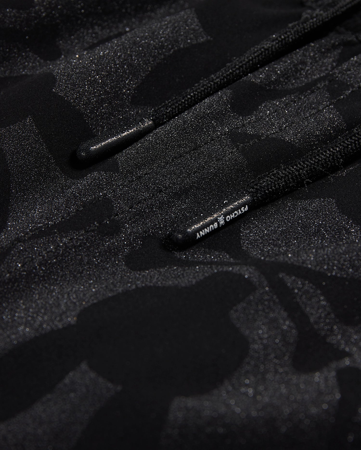 Close-up of black polyester mechanical stretch fabric with a subtle camo pattern and glossy accents, featuring drawstrings with visible Psycho Bunny branding.