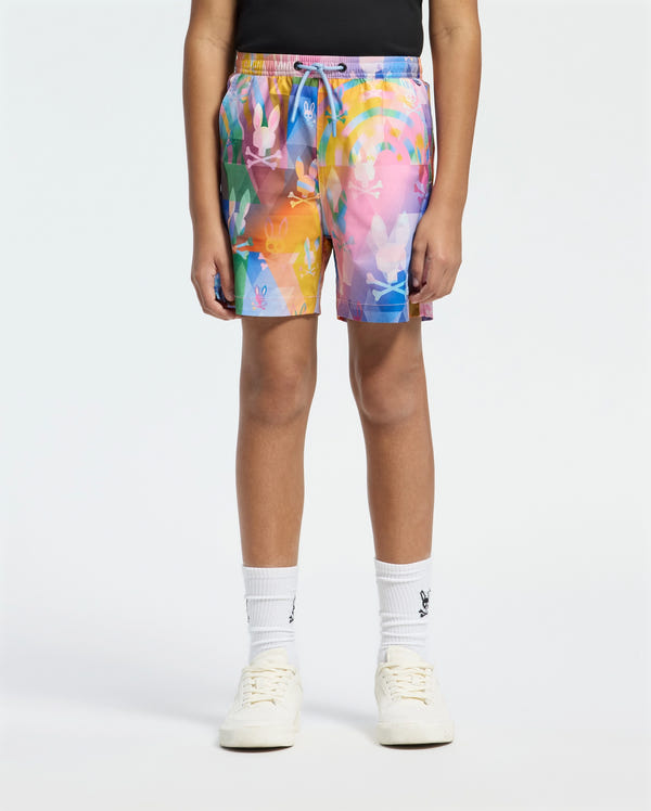 A person in Psycho Bunny KIDS MILOS PRINTED SWIM TRUNK - B0W446F200, adorned in an abstract pattern, pairs them with a black top, white socks, and sneakers against a plain white background. The quick-dry fabric makes it ideal for vacation style.