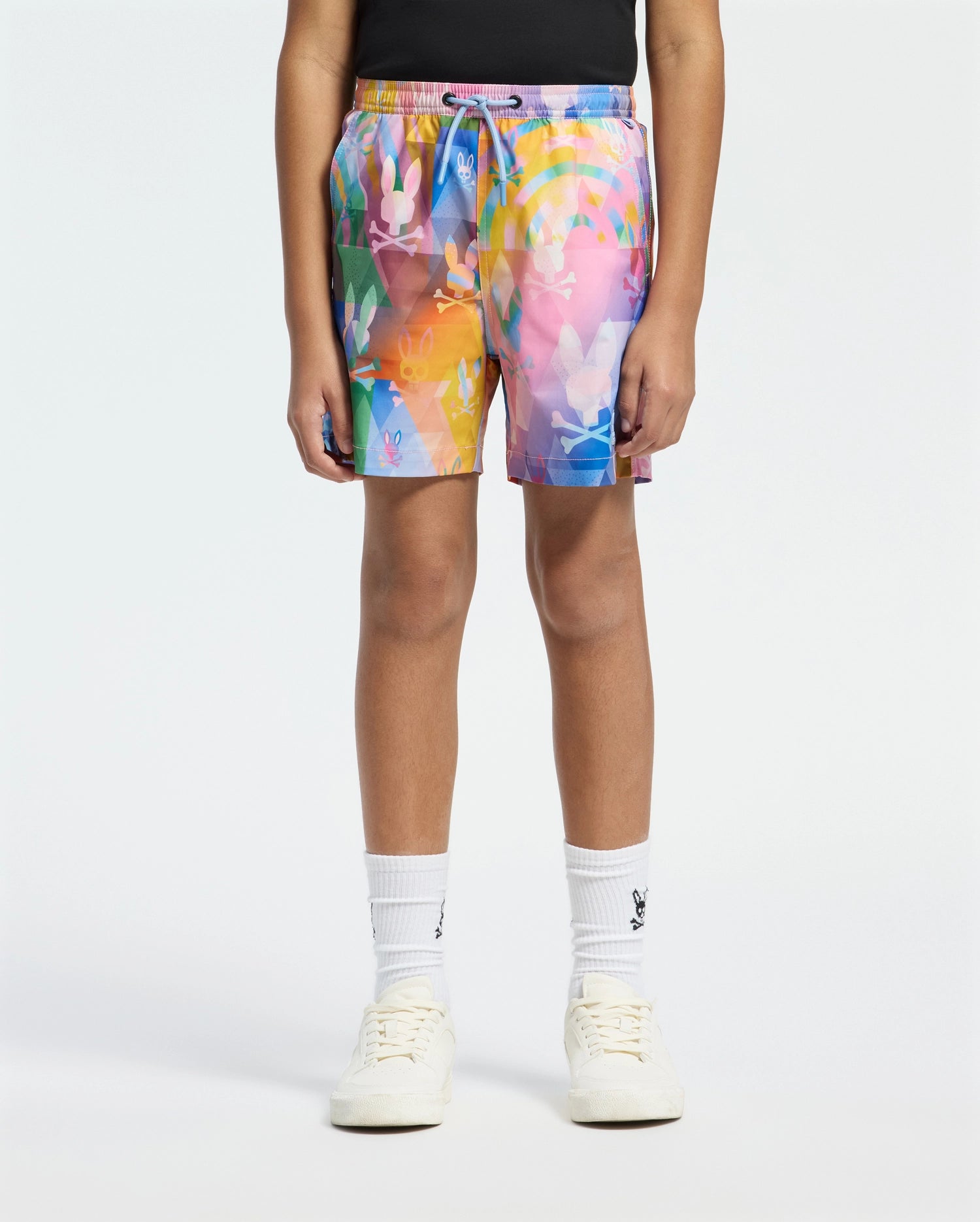 A person in Psycho Bunny KIDS MILOS PRINTED SWIM TRUNK - B0W446F200, adorned in an abstract pattern, pairs them with a black top, white socks, and sneakers against a plain white background. The quick-dry fabric makes it ideal for vacation style.