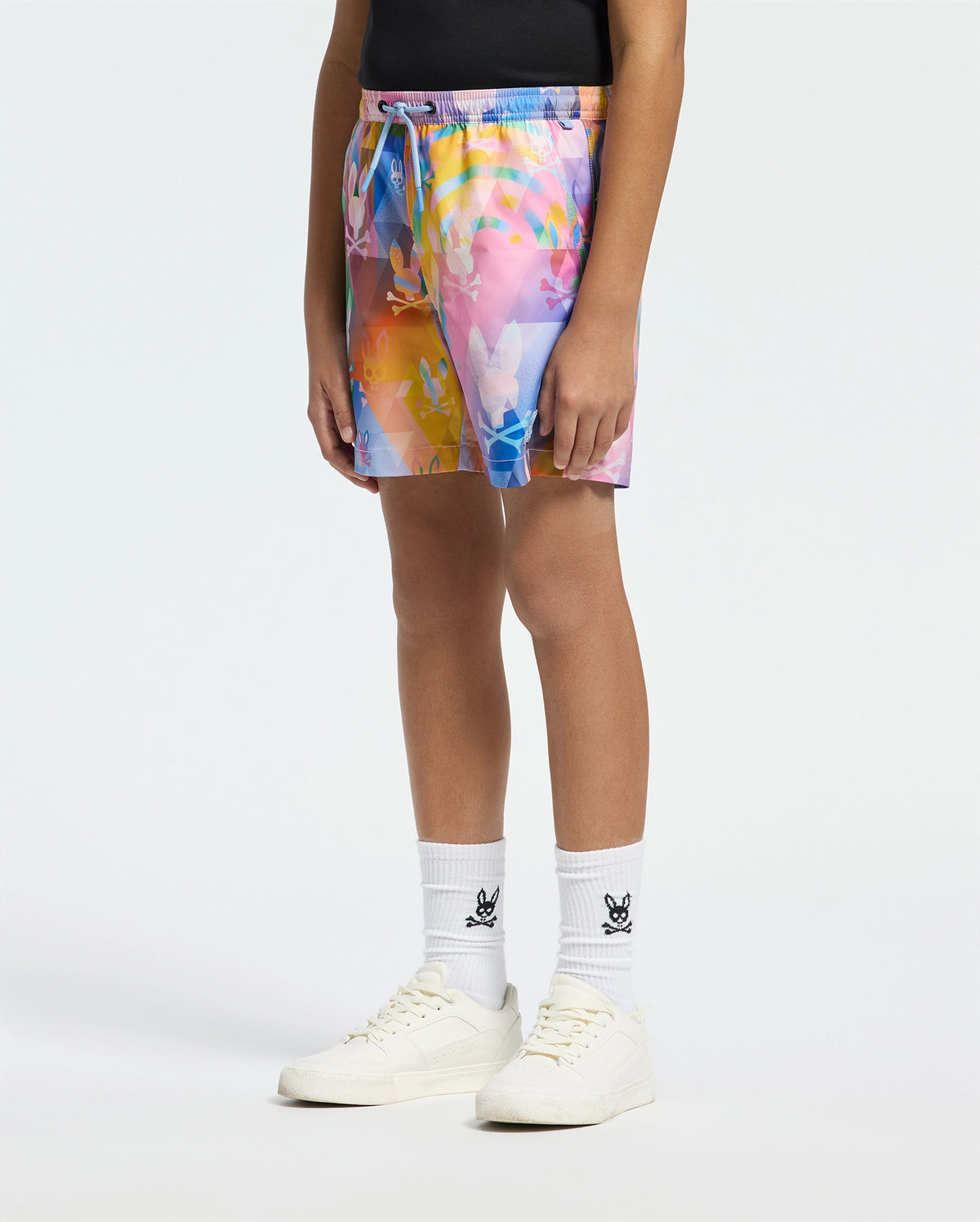 A person flaunts their vacation style in colorful abstract-patterned Psycho Bunny Kids Milos Printed Swim Trunk - B0W446F200. They pair the vibrant shorts with white socks featuring a black logo and classic white sneakers, with the quick-dry fabric enhancing their bold look against a plain white background.
