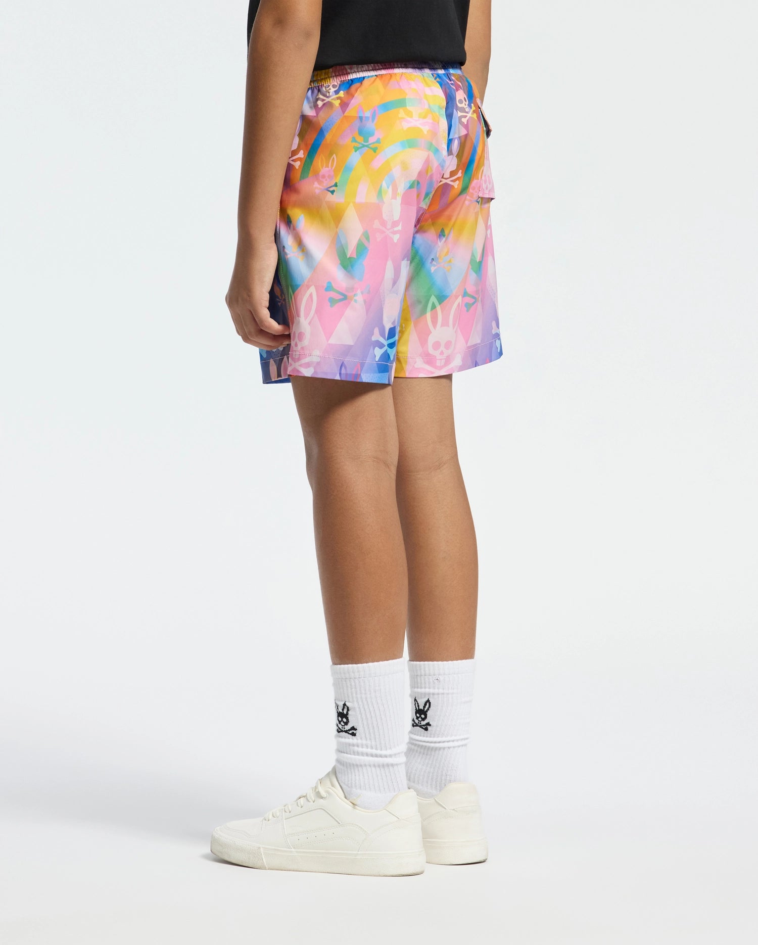 A person wears Psycho Bunny KIDS MILOS PRINTED SWIM TRUNK (B0W446F200) featuring a colorful, graffiti-like pattern, paired with white socks with black logo designs and white sneakers. The quick-dry fabric is ideal for vacations, all set against a simple white background to highlight the outfit.