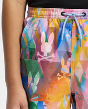 A person wears KIDS MILOS PRINTED SWIM TRUNK by Psycho Bunny, featuring abstract bunnies and skulls in vibrant pink, blue, and yellow. Ideal for vacation with its quick-dry fabric, the trunks are secured at the waist by blue drawstrings as their hand rests by their side.