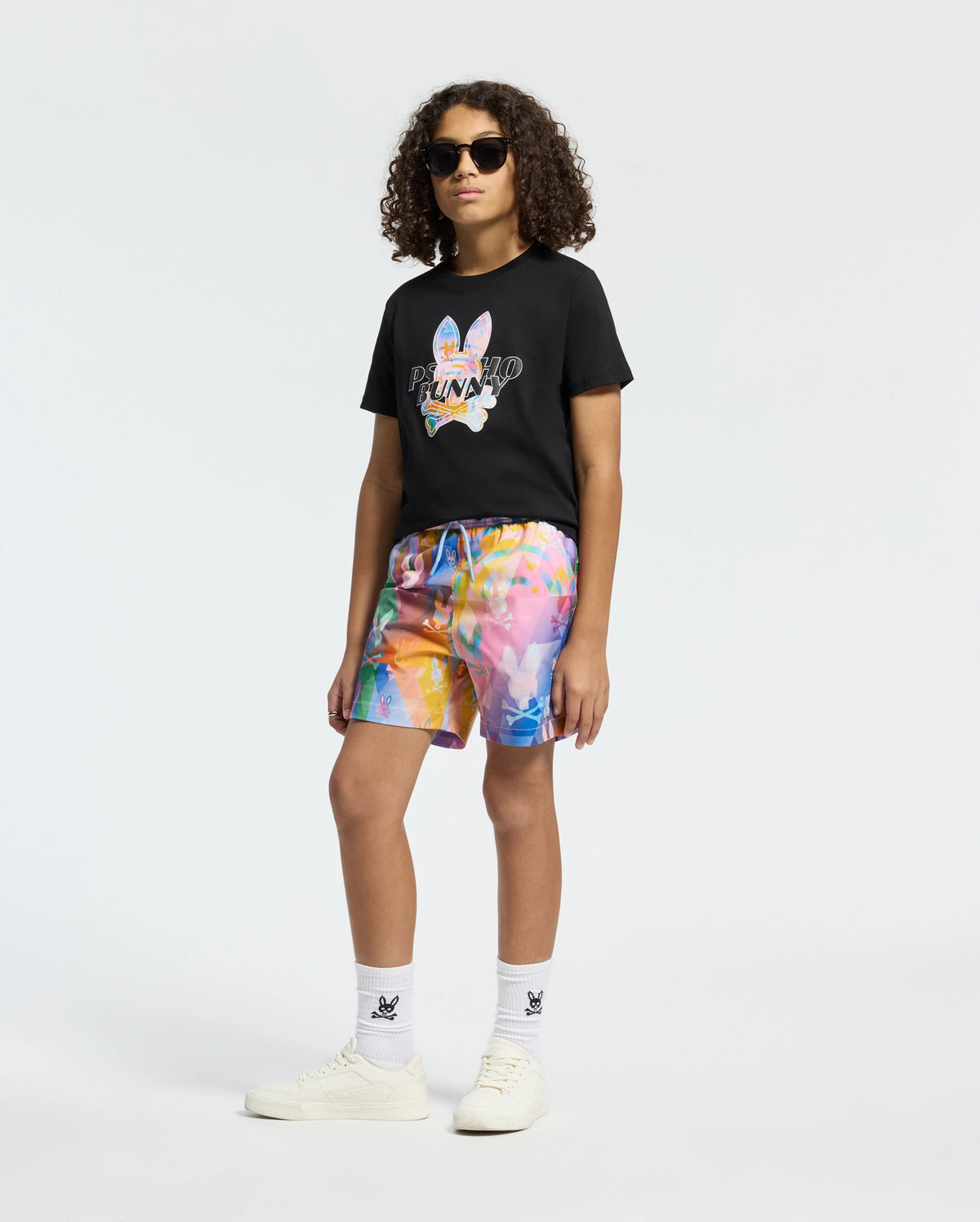 A person with curly hair and sunglasses exudes effortless vacation style in a black graphic tee and striking KIDS MILOS PRINTED SWIM TRUNK by Psycho Bunny. Completing the look with white socks and sneakers, they stand confidently against a plain white background.