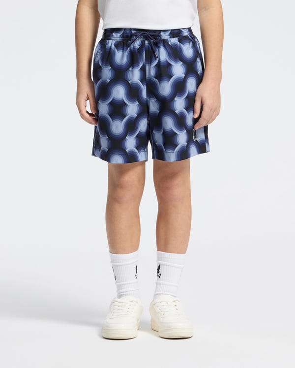 A person in a white shirt, Psycho Bunny KIDS PAROS PRINTED SWIM TRUNK - B0W450F200 with a blue geo-pattern and bold wavy design for mechanical stretch, wears white socks and sneakers. They stand against a plain background with hands in their pockets.