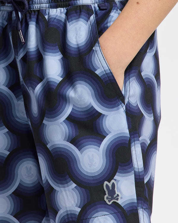 A close-up of a person wearing KIDS PAROS PRINTED SWIM TRUNK by Psycho Bunny features geo-patterned swim shorts with a blue and black circular design, showing the small bunny logo near the hem as one hand rests in the pocket, enjoying comfort from mechanical stretch.