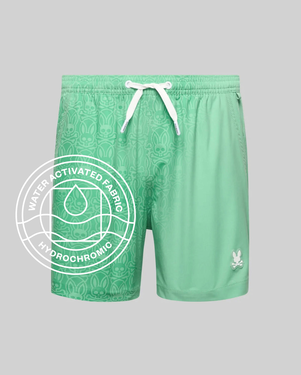 Kids Samos Hydrochromic Printed Swim Trunk in mint green, featuring a relaxed fit, white drawstrings, and a small white Psycho Bunny skull logo on the lower right leg. The smooth fabric reveals a tonal bunny skull pattern when wet.