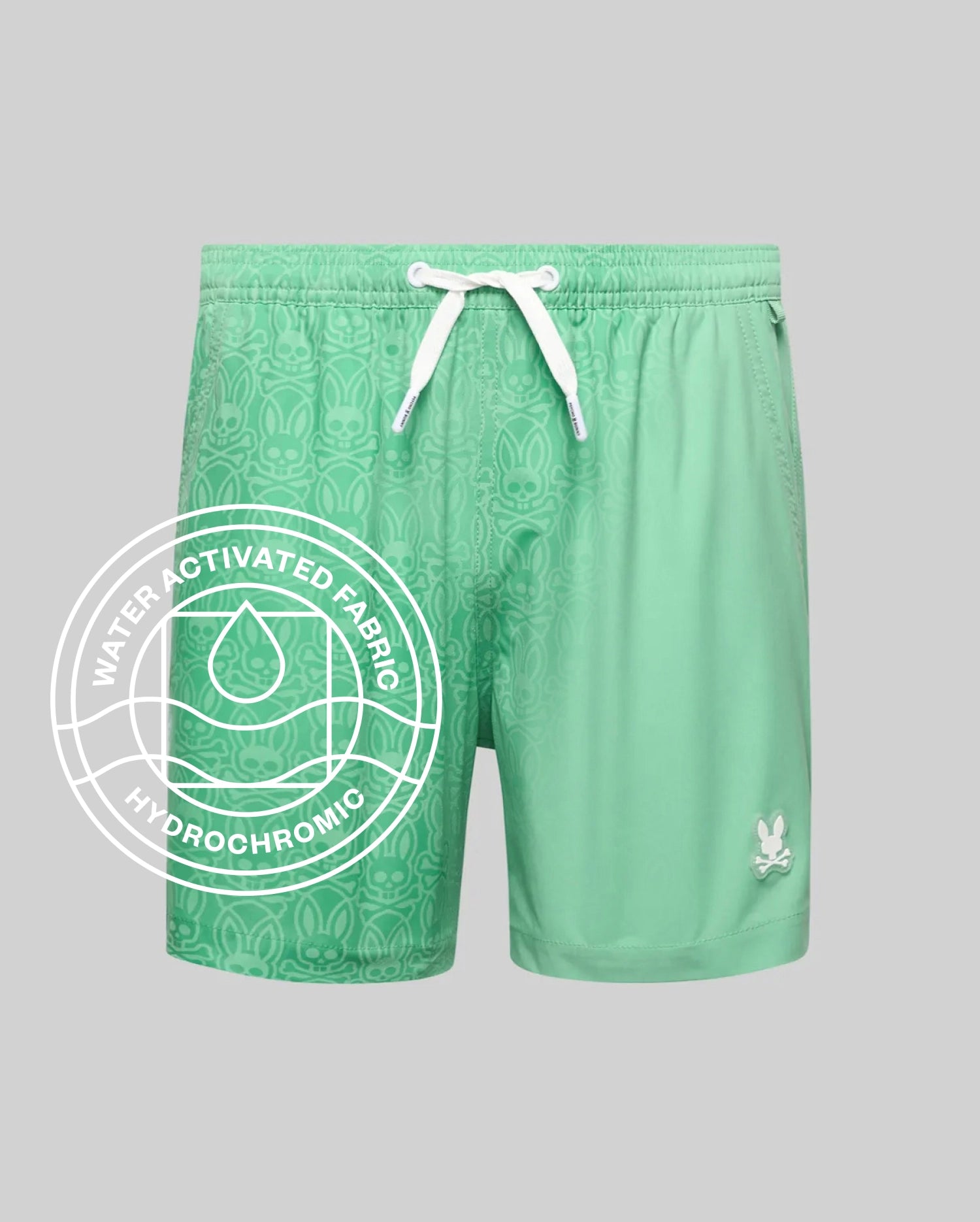Kids Samos Hydrochromic Printed Swim Trunk in mint green, featuring a relaxed fit, white drawstrings, and a small white Psycho Bunny skull logo on the lower right leg. The smooth fabric reveals a tonal bunny skull pattern when wet.